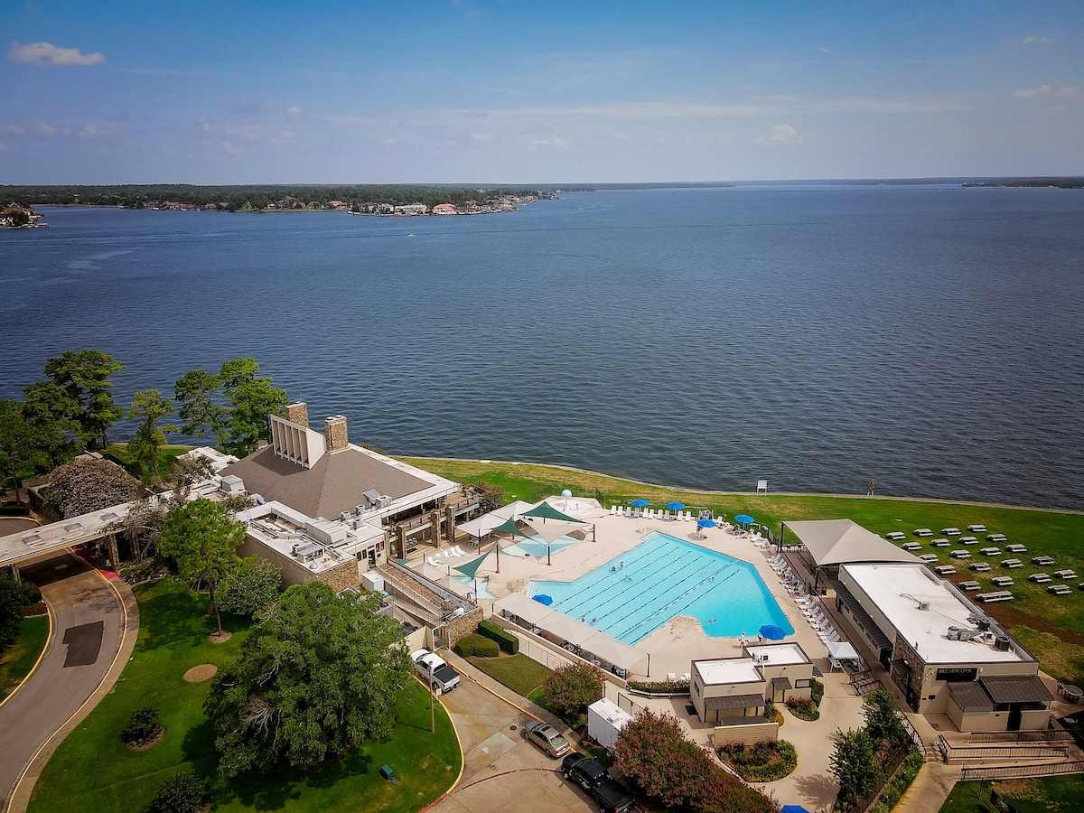 An aerial view showcases a large swimming pool adjacent to the lake, surrounded by well-maintained lawns and lounge chairs. The tranquil water reflects the sky, offering a serene atmosphere. The nearby building features outdoor seating areas and is set amidst lush trees.
