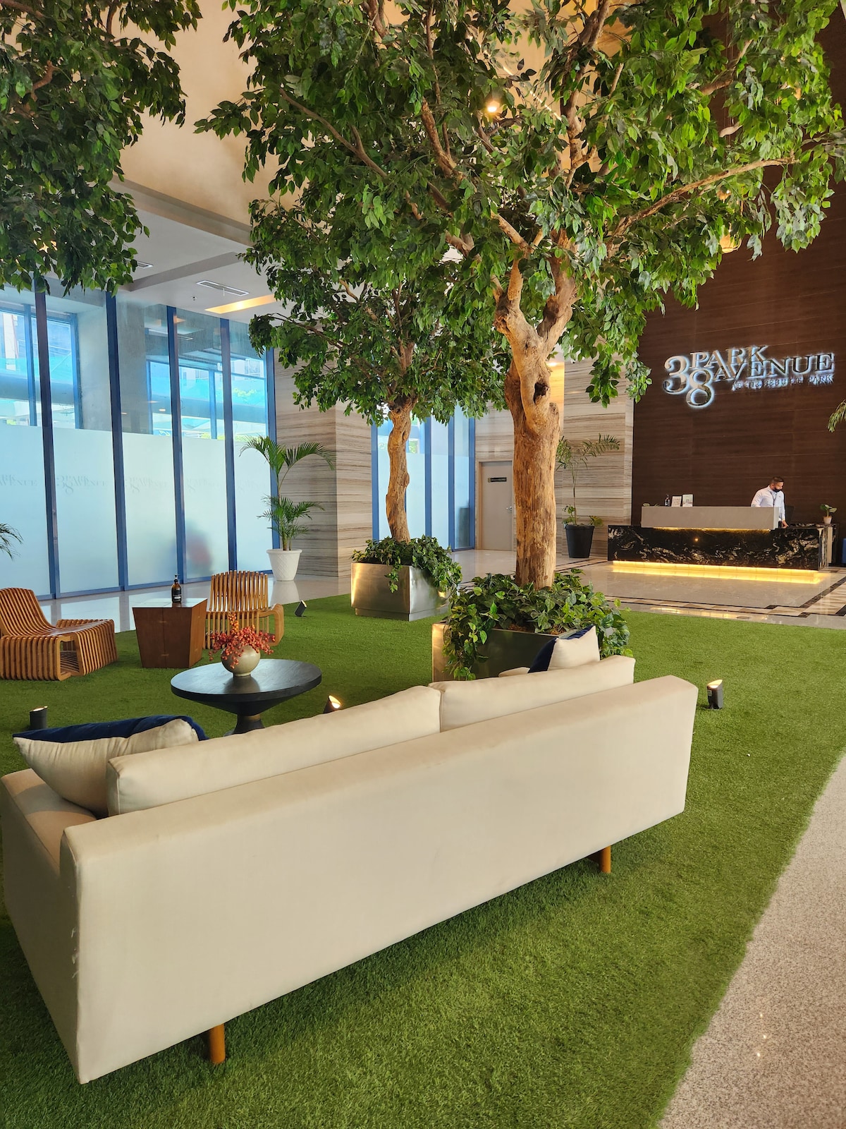 An inviting lobby area features a light-colored sofa and modern seating arrangements surrounded by greenery. Tall trees are strategically placed among the furniture, and a sleek reception desk is visible in the background, providing a welcoming ambiance.