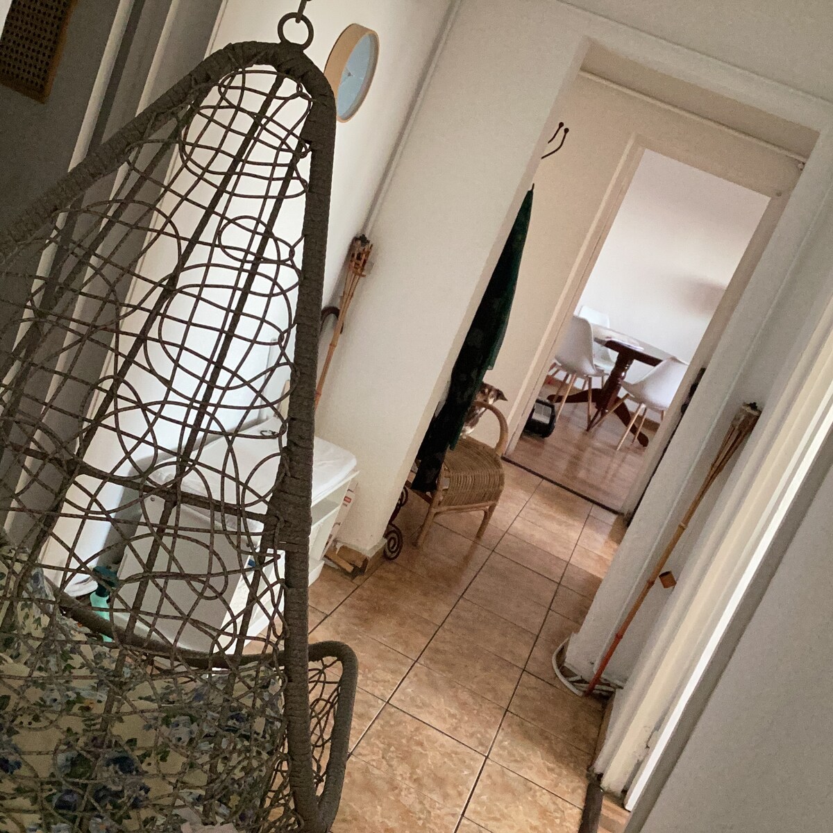 A hallway is showcased, featuring a unique hanging chair with a patterned cushion. Light-colored walls create a bright atmosphere, while tile flooring extends down the corridor. At the end of the hall, a dining area is subtly visible, complemented by a selection of chairs.