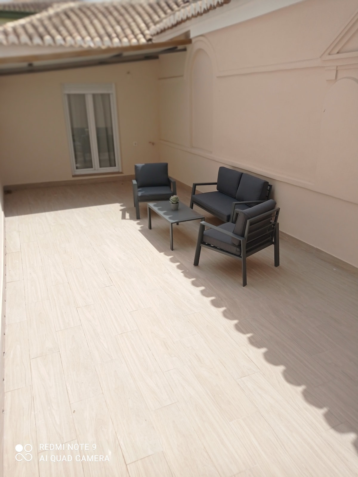 A spacious terrace features modern seating with two chairs and a coffee table, all set on a light tiled floor. Natural light enters through a sliding glass door, illuminating the area. A soft neutral color palette creates a calm outdoor setting.