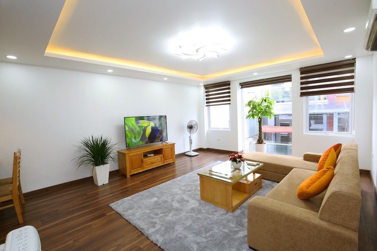 A modern living area features an inviting sofa with orange accent pillows and a glass coffee table at its center. Natural light filters through two windows adorned with wooden blinds. A flat-screen TV and decorative plant enhance the space, complemented by warm lighting from the ceiling.