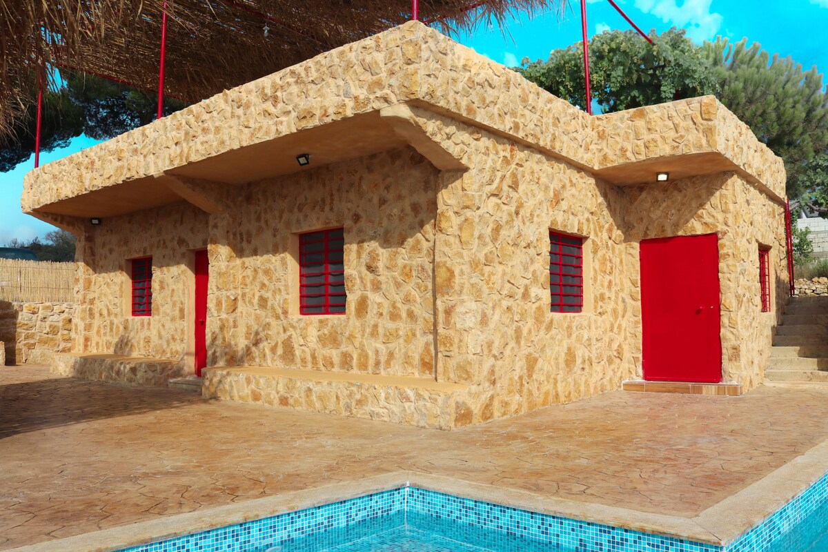A single-story, stone house features textured walls and vibrant red doors and window frames. The structure is surrounded by a spacious patio area, with a private pool visible in the foreground, offering a relaxed outdoor atmosphere.