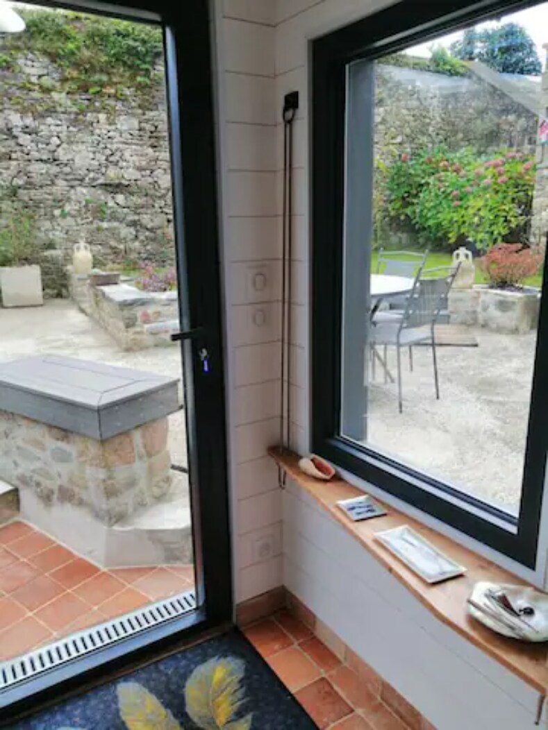 A view of a cozy interior space is captured, featuring large windows that overlook a private garden area. A stone wall provides a rustic backdrop, while a small table and chairs can be seen outside, accompanied by greenery and ornamental elements in the garden.