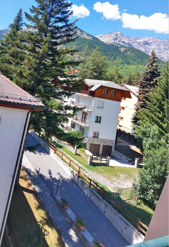 A view reveals nearby buildings framed by tall trees, with mountains visible in the background. A quiet path is lined with planters, leading to an open green area. The scene reflects a tranquil setting surrounded by nature in a well-maintained neighborhood.