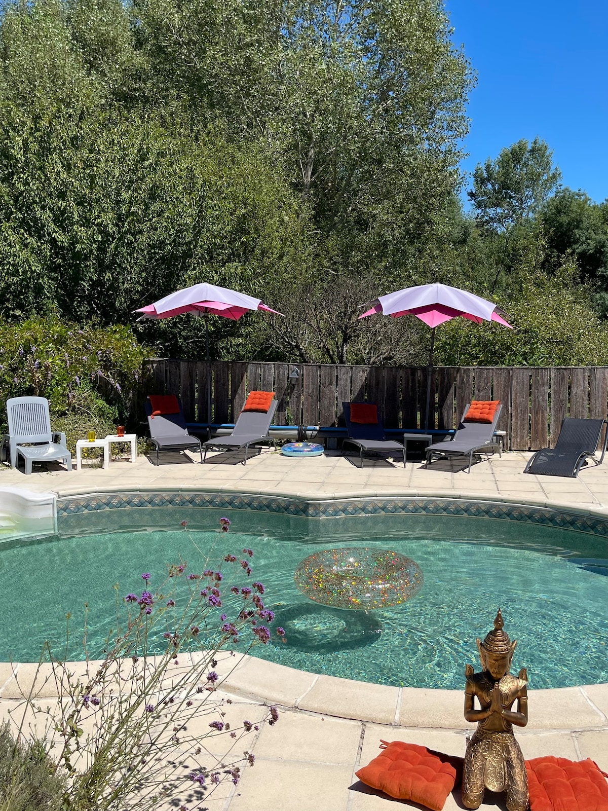 An inviting pool area is surrounded by lush greenery. Colorful lounge chairs are arranged under pink and white umbrellas. A decorative glass float rests in the water, while a small statue adds a serene touch to the outdoor space.
