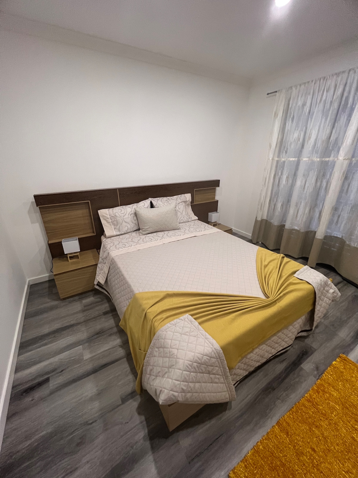 A well-appointed bedroom is featured, showcasing a spacious bed with a light-colored quilt draped across the foot. The bed is flanked by wooden nightstands, and a sheer curtain allows natural light to filter through, illuminating the sleek flooring and adding warmth to the space.
