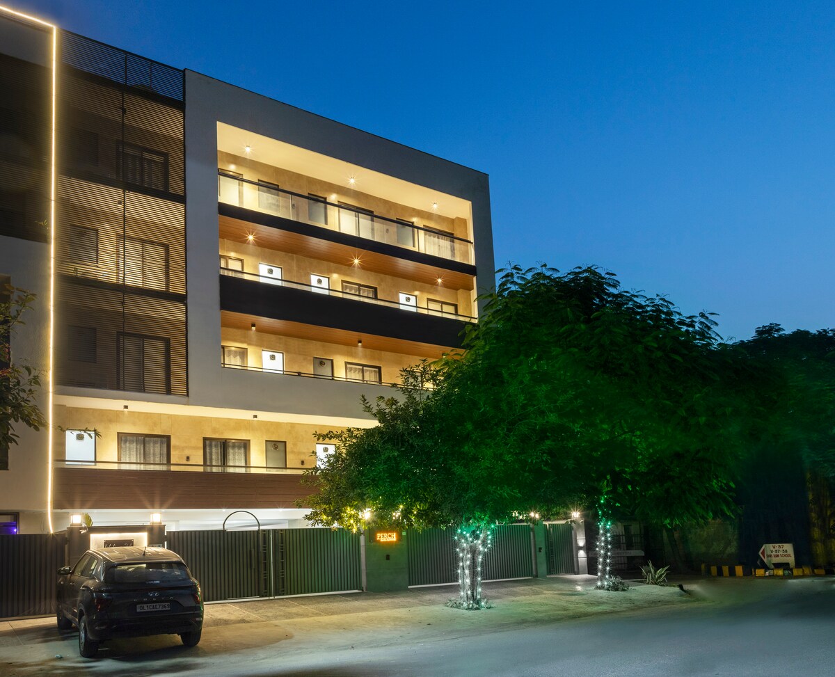 The three-story building is illuminated with soft lighting, enhancing its modern design. Balconies are featured on each floor, surrounded by lush greenery. A vehicle is parked in front, providing a sense of accessibility and welcoming atmosphere.