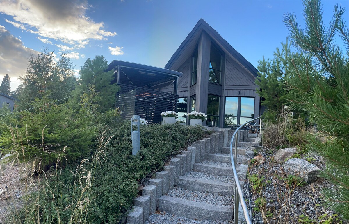 The exterior view of a modern villa is showcased, featuring large glass windows and a sloping roof. A landscaped pathway of stone steps leads to the entrance, surrounded by greenery and flower beds. A covered outdoor seating area is visible to the left.