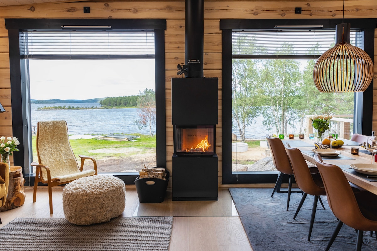 A cozy interior space features a stylish wood-burning stove as a focal point, surrounded by large windows offering views of the lake. A round dining table with several chairs is visible, alongside a comfortable seating area furnished with a soft textured chair.