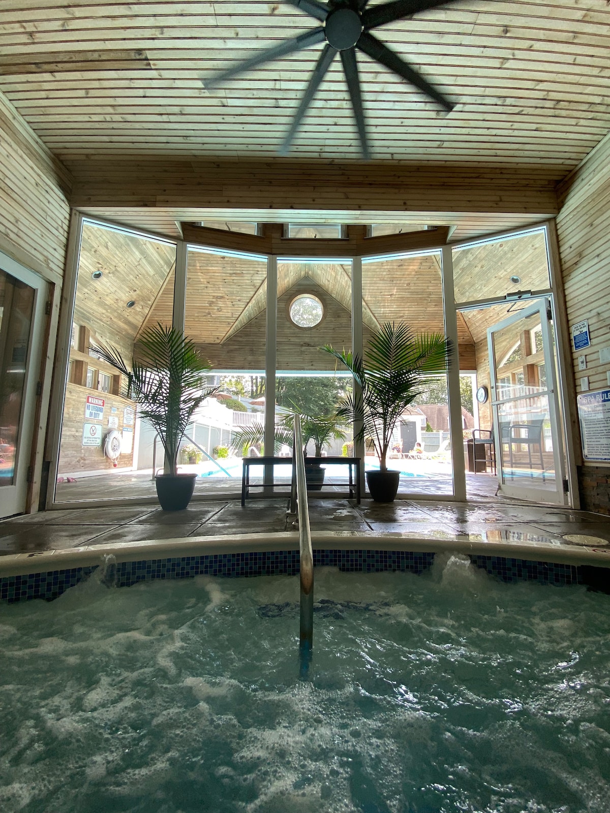 An indoor hot tub is visible, surrounded by wooden walls and a high ceiling. Large glass doors provide a view of the outdoor pool area, enhanced by lush potted plants on either side. A circular window allows natural light to fill the space.