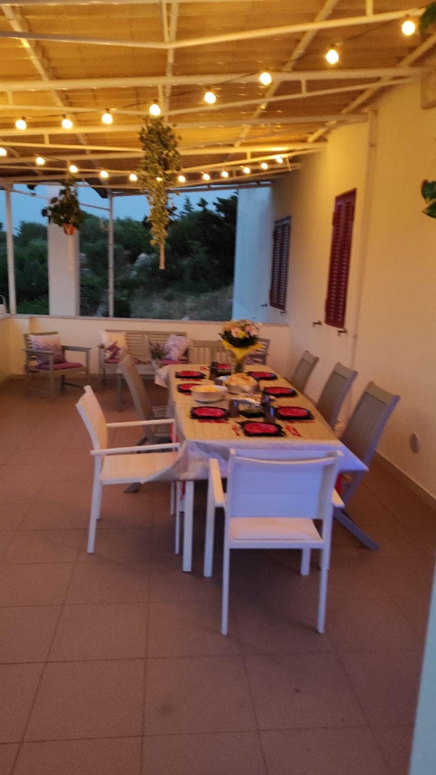 An outdoor dining area features a long table set for guests, surrounded by modern chairs. Soft string lights hang above, adding a gentle warmth. The area is partially enclosed with large windows, providing views of the surrounding greenery.