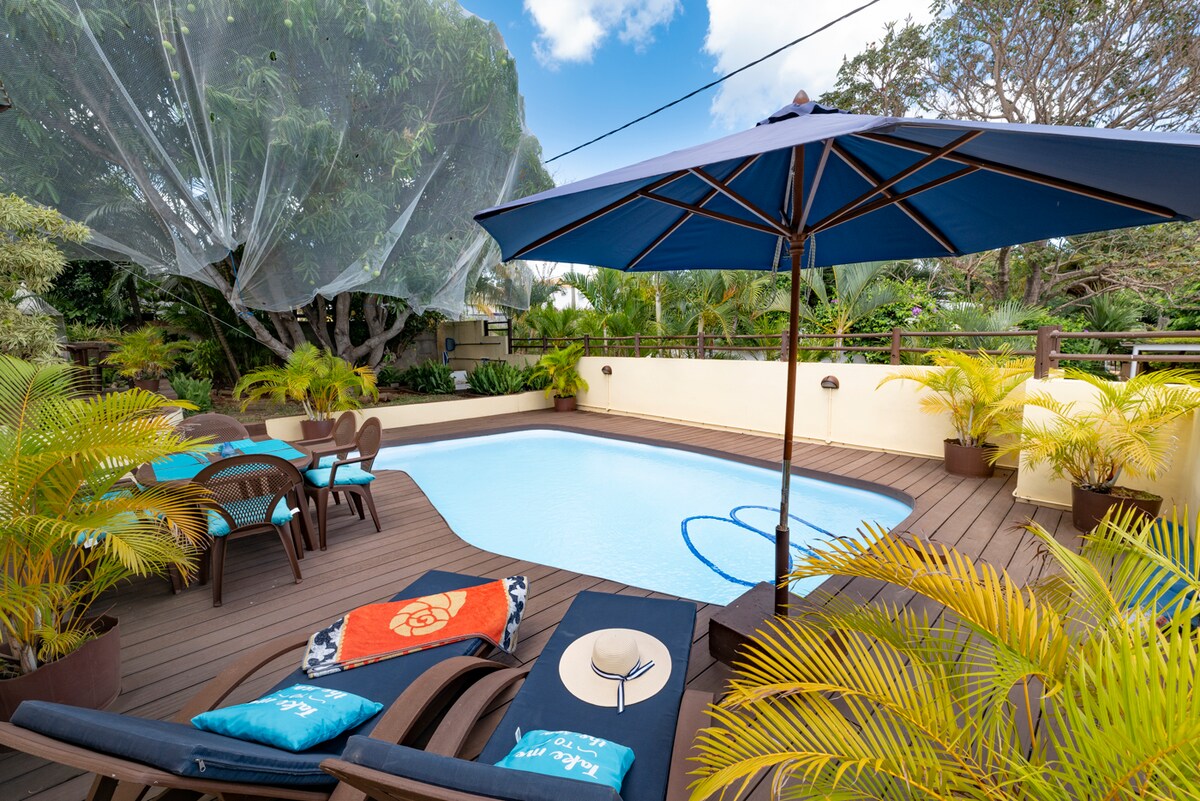A serene outdoor space is highlighted by a refreshing pool surrounded by lush greenery. Sun loungers with colorful towels provide comfort, while an umbrella offers shade. A nearby dining area with chairs is positioned for relaxation and enjoyment in this private garden setting.