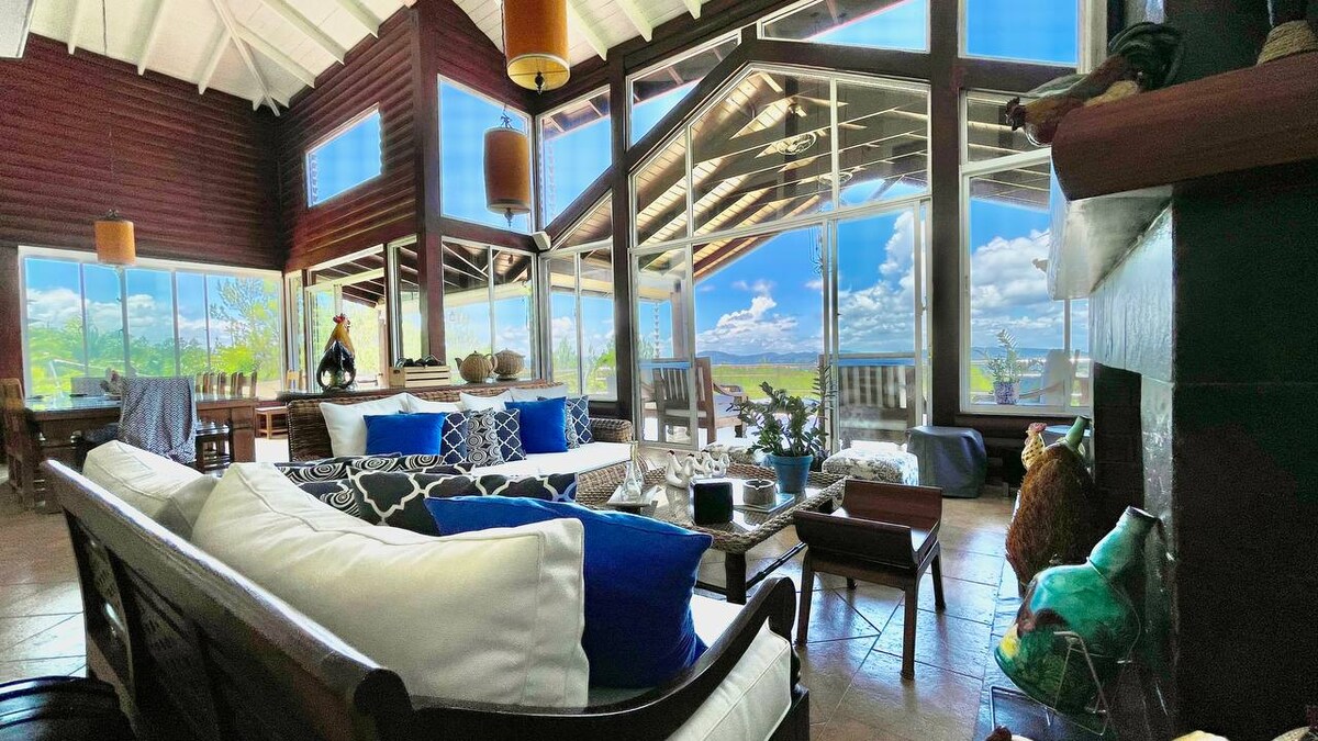 A spacious living area is centered around comfortable seating, featuring a large sofa adorned with blue and white cushions. The room is filled with natural light from expansive glass windows that provide scenic views of the surrounding mountains and outdoor space.
