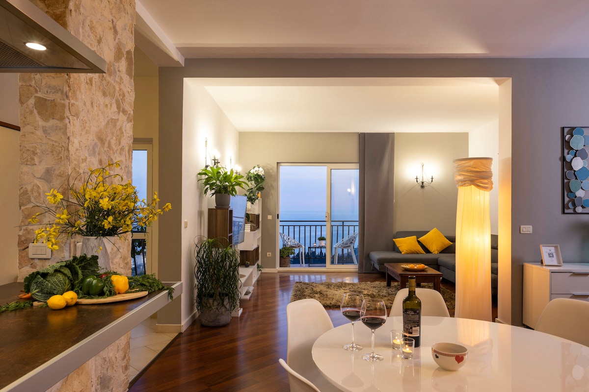 A spacious living area features large windows framing a sea view, enhanced by soft lighting. A dining table is set with wine and fresh produce. A comfortable seating area includes a couch with decorative cushions, and vibrant plants add a touch of greenery.