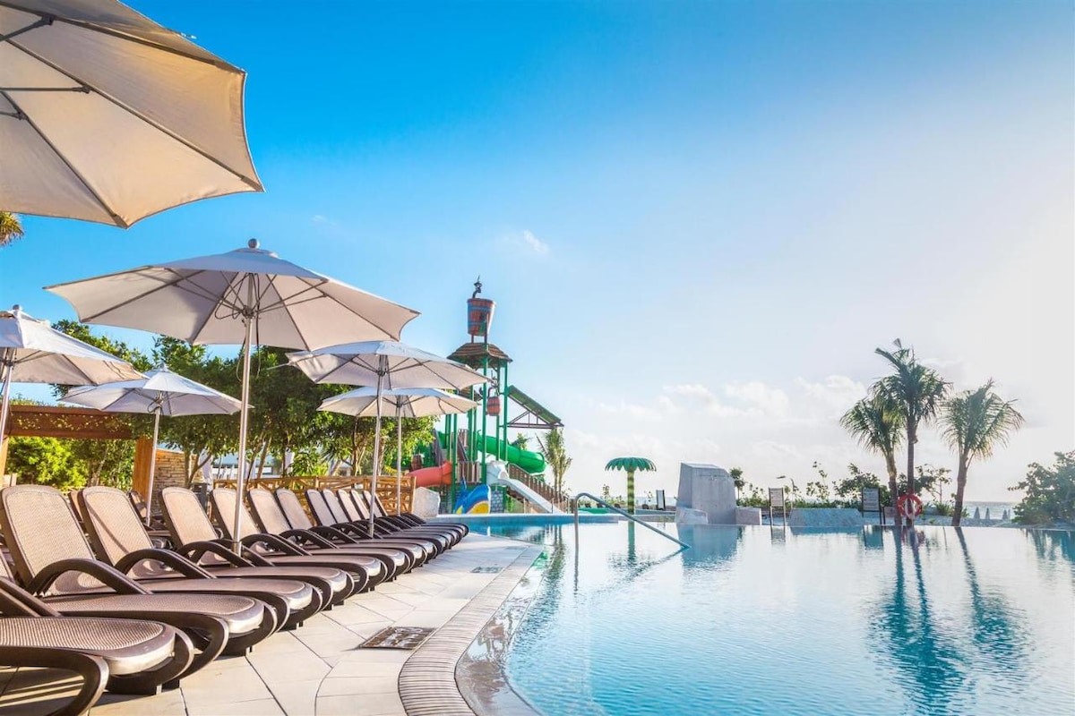 A shimmering outdoor swimming pool is surrounded by lounge chairs, each paired with a shade-providing umbrella. In the background, a colorful water slide adds a playful element against a clear blue sky with a few scattered clouds.