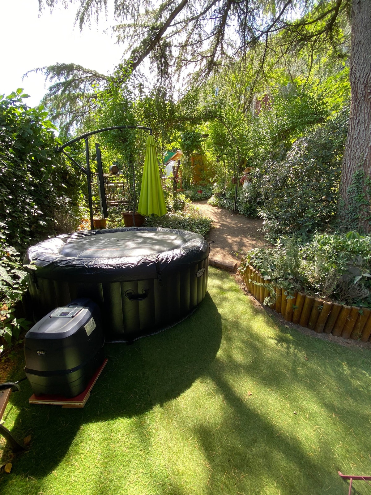 A private jacuzzi is positioned on a grassy area surrounded by lush greenery. A pathway lined with plants leads away from the jacuzzi, suggesting a serene garden setting. Sunlight filters through the trees, enhancing the tranquil environment.
