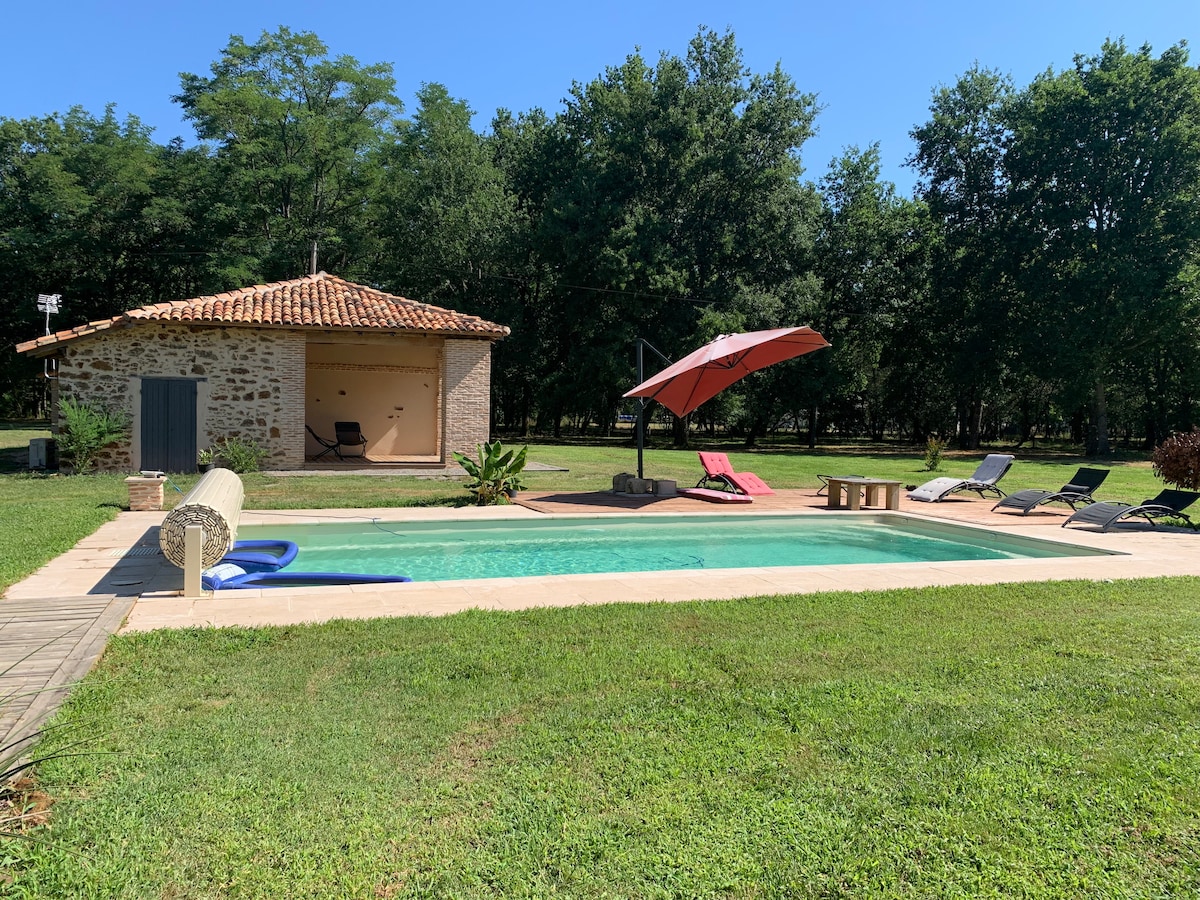 A refreshing outdoor pool is surrounded by a spacious grassy area, with comfortable lounge chairs positioned for relaxation. A shaded area with an umbrella offers respite from the sun, complemented by a charming stone house in the background, featuring a traditional tiled roof.