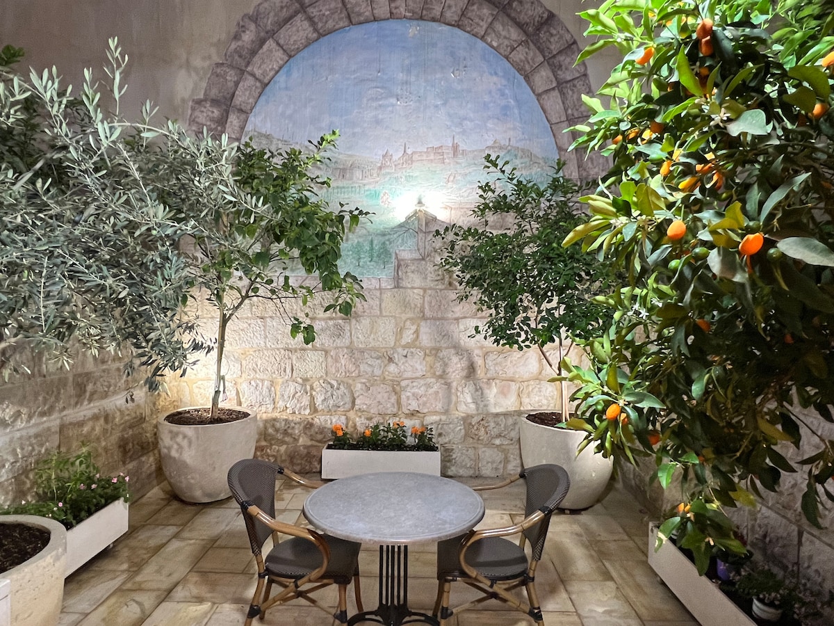 A serene garden area features a round stone table surrounded by four cushioned chairs. Lush greenery, including olive trees and citrus plants in white planters, adds a natural touch against a stone wall painted with a scenic mural.