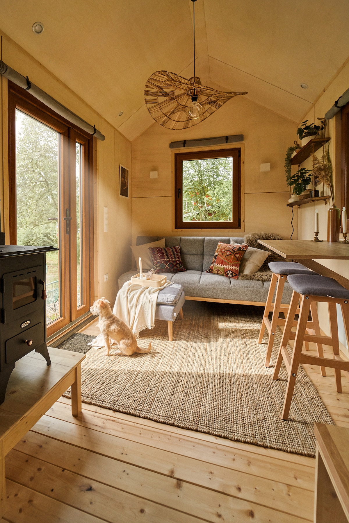 A cozy living area features a comfortable gray sofa adorned with decorative pillows. Natural light enters through a large window, illuminating wooden walls and a woven area rug. A small dining space with two stools is positioned beside a decorative stove, enhancing the inviting environment.