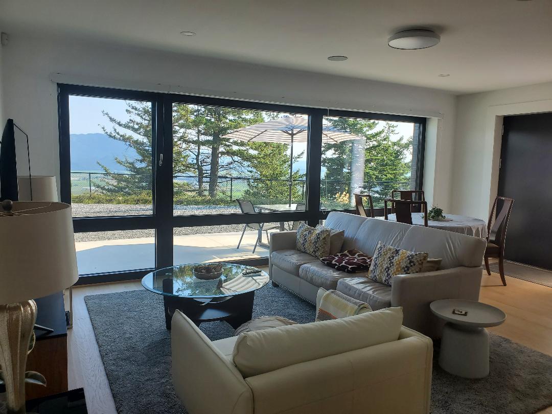 A spacious living area features large glass windows, showcasing views of the surrounding nature. A light-colored sofa is arranged with decorative cushions, accompanied by a round coffee table. A dining table is set in the corner, with sunlight filtering through the windows.