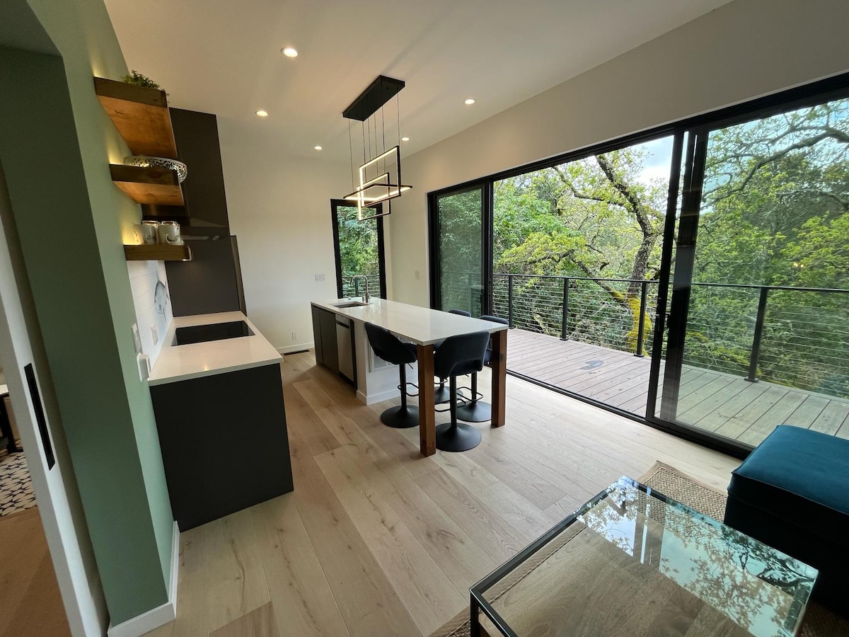 A modern kitchen is featured with sleek cabinets and a white countertop. A dining table with four black chairs is positioned beneath a contemporary light fixture. Expansive glass doors lead to an outdoor deck surrounded by lush greenery, enhancing the connection to nature.