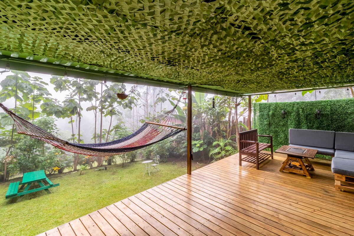 A spacious outdoor area features a wooden deck with a seating arrangement, including a bench and a low table. A colorful hammock is suspended for relaxation, surrounded by lush greenery. The area is shaded by a patterned canopy, providing a serene atmosphere.