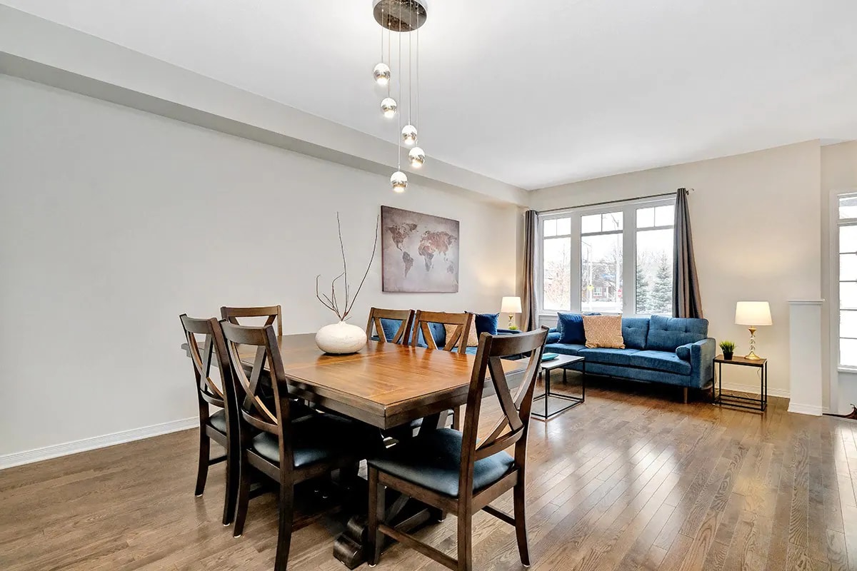 A modern dining area features a large wooden table surrounded by six dark chairs. A cozy blue couch is positioned nearby, complemented by a lamp and window treatments. A world map artwork is hung on the wall, contributing to the inviting ambiance.