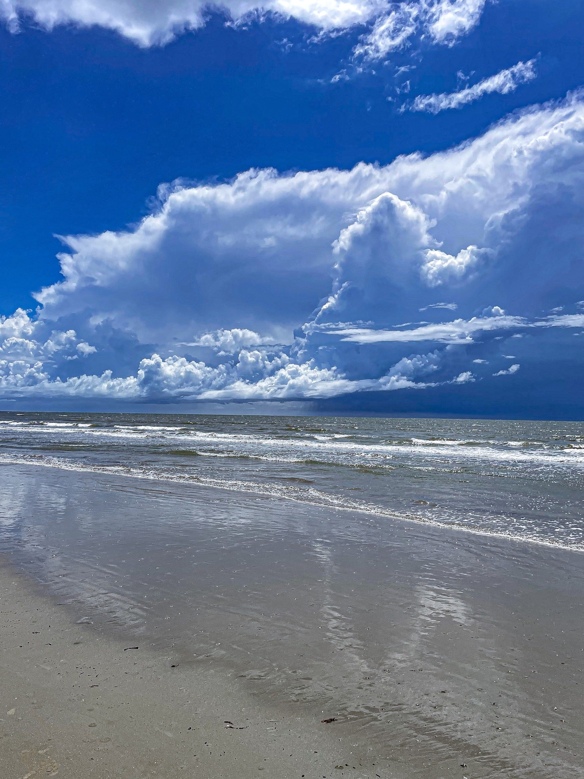 The image captures a serene beach scene under a vibrant blue sky, dotted with large, fluffy clouds. The water gently laps at the shore, reflecting the cloud formations, while the sandy beach remains smooth and unruffled, inviting a peaceful stroll along the coast.