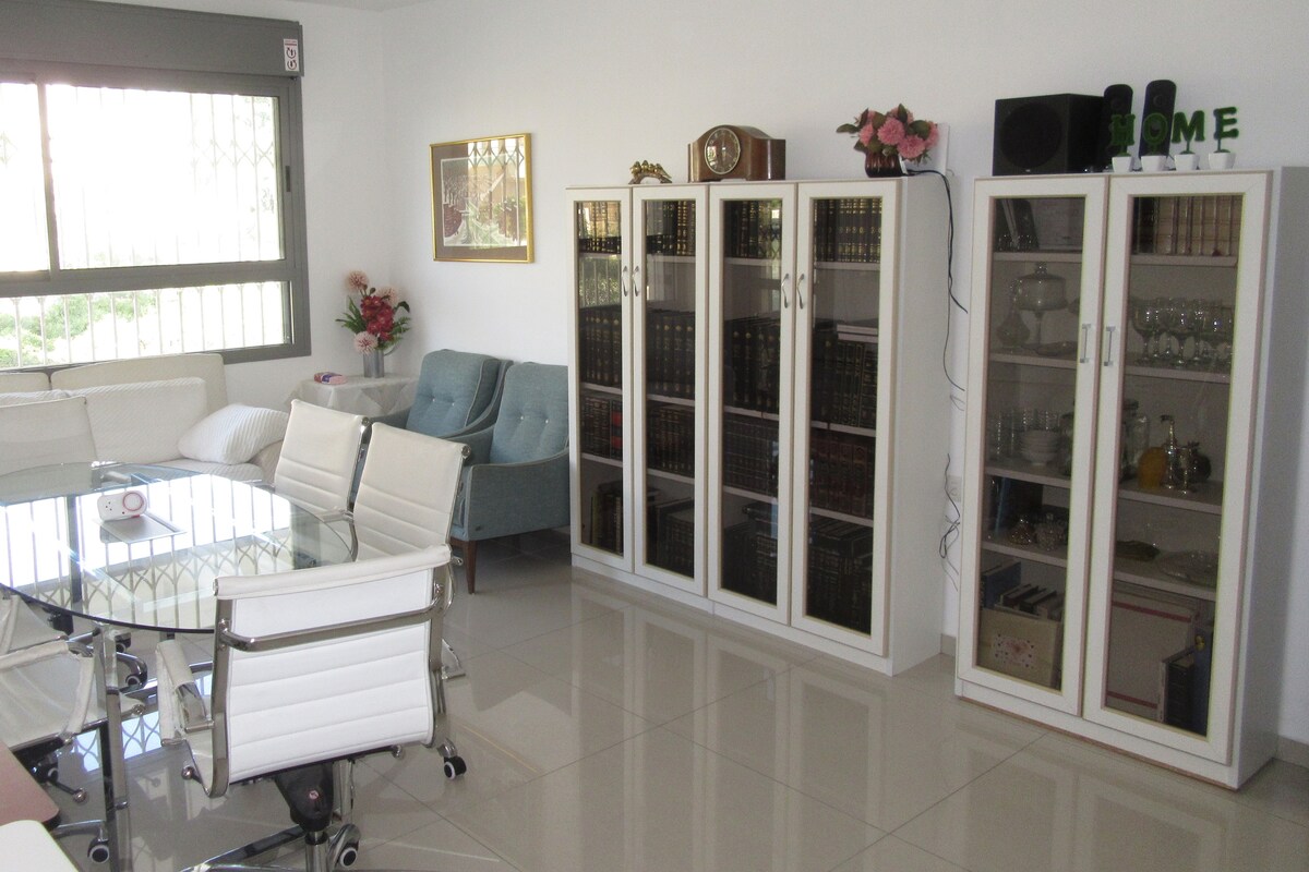 A bright living area features a glass dining table surrounded by white chairs. Nearby, two white display cabinets showcase books and decorative items. A comfortable seating area includes a light blue armchair. Natural light flows in through a large window with a view of the outdoors.