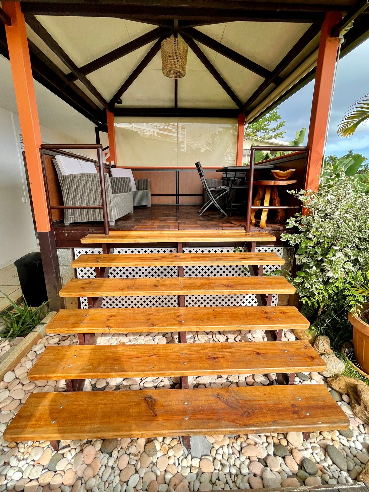 A wooden staircase leads up to a covered outdoor seating area. The space features a comfortable couch and a dining table, with lush greenery surrounding the entrance. The roof extends gracefully to provide shade, enhancing the inviting atmosphere.