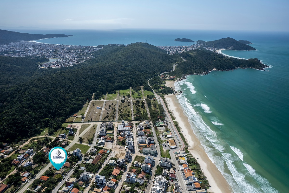An aerial view captures the coastline with its gentle waves and sandy shores, bordered by lush green hills. The image shows the contrast between urban development and natural scenery, highlighting the beautiful azure waters and the expansive beach area, ideal for relaxation and enjoyment.