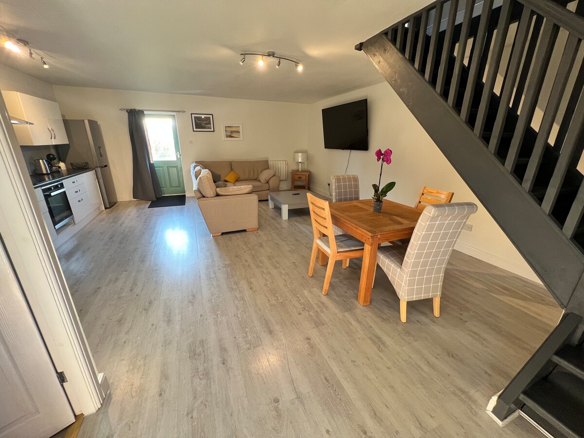 An open-plan living area is featured with a spacious seating arrangement, a dining table with four chairs, and a modern kitchen. A large 75" Smart TV is mounted on the wall, while light enters through a door leading to the outside. A staircase is visible in the corner.