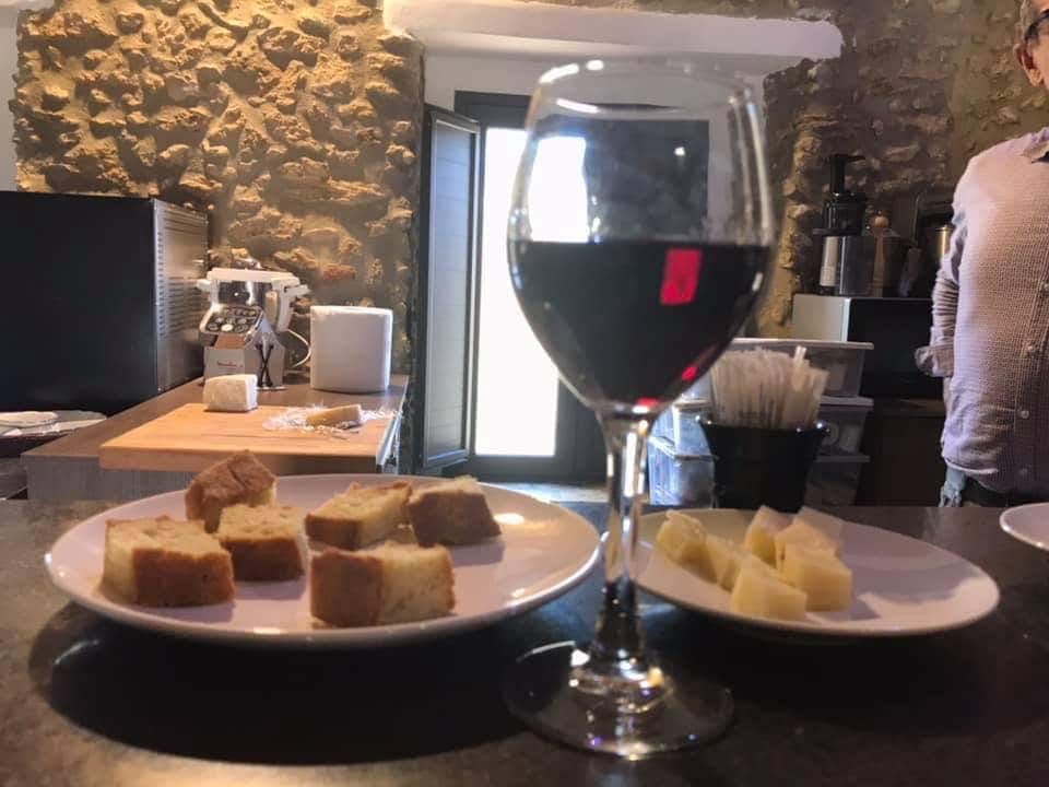 A glass of red wine is positioned beside two white plates. One plate holds cubed pieces of bread, while the other contains slices of cheese. The rustic stone wall in the background is complemented by an open door, revealing a glimpse of the outdoor light.