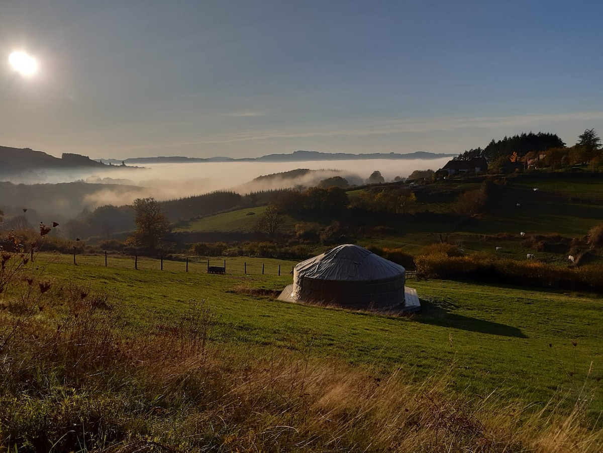 A traditional Mongolian yurt is positioned on a grassy slope, surrounded by a lush landscape. The morning sun casts a warm glow as mist blankets the valley below, revealing hills in the distance. The serene scene reflects a peaceful and natural setting.