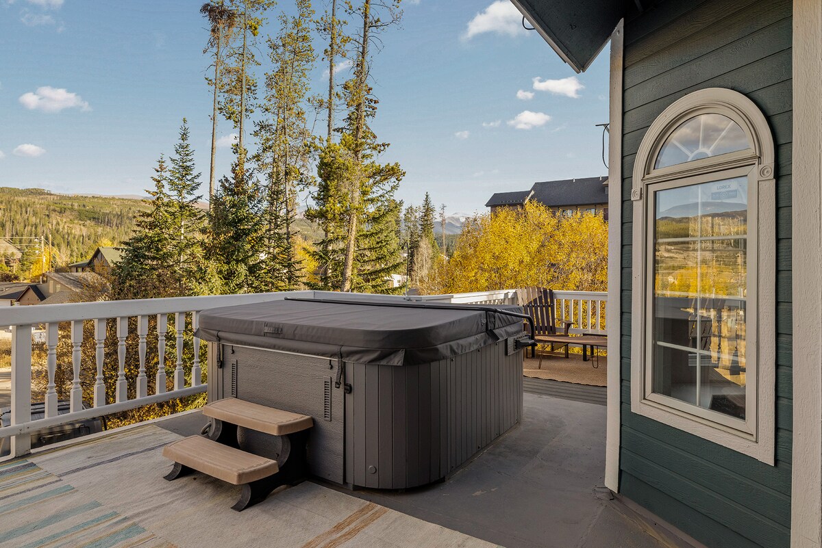 A private hot tub is positioned on a spacious deck, surrounded by trees with golden foliage. A nearby bench offers seating with expansive views of the mountainous landscape. Large windows provide natural light to the room, enhancing the serene outdoor ambiance.