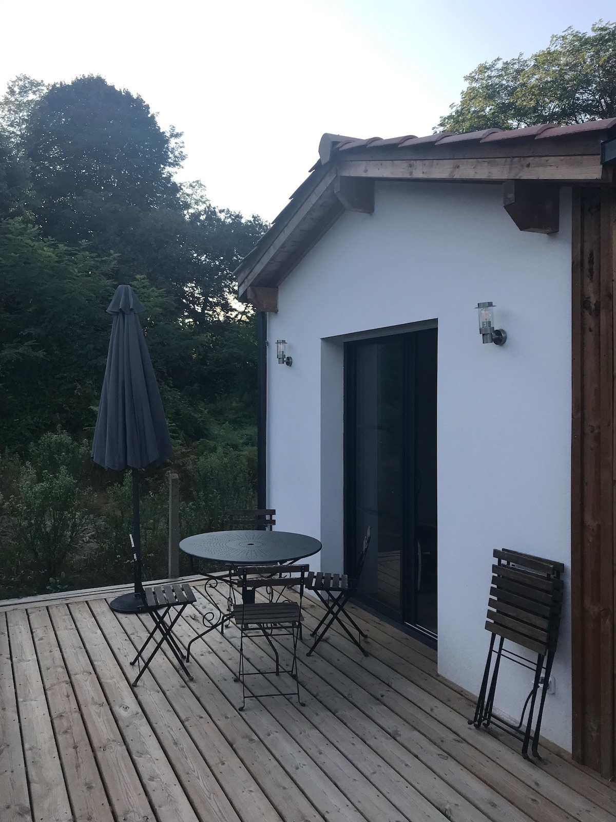 A private terrace features a round table surrounded by four folding chairs. A large umbrella provides shade, while the wooden deck is bordered by lush greenery. The sliding glass doors lead into the living space, enhancing an outdoor relaxation area.
