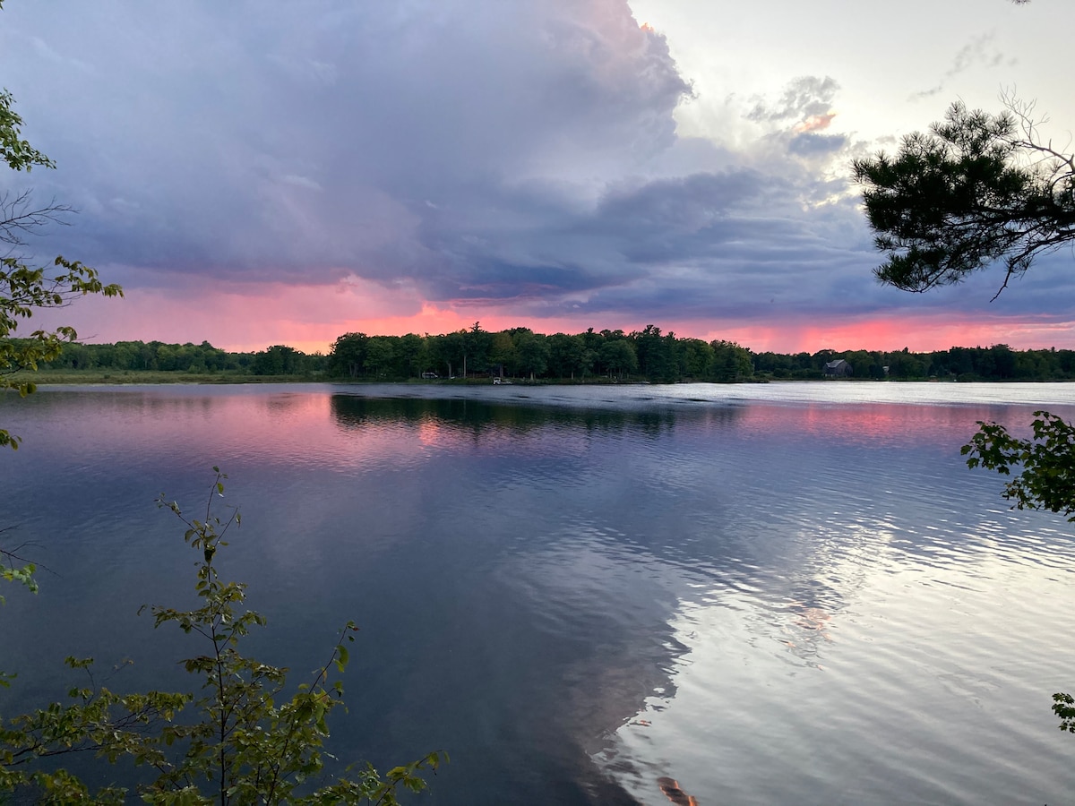 A serene lake scene is captured, with gentle ripples reflecting the colorful sky. Shades of pink and purple are visible amidst the clouds, while lush greenery surrounds the water's edge. The calm surface of the lake creates a peaceful ambiance, perfect for relaxation.