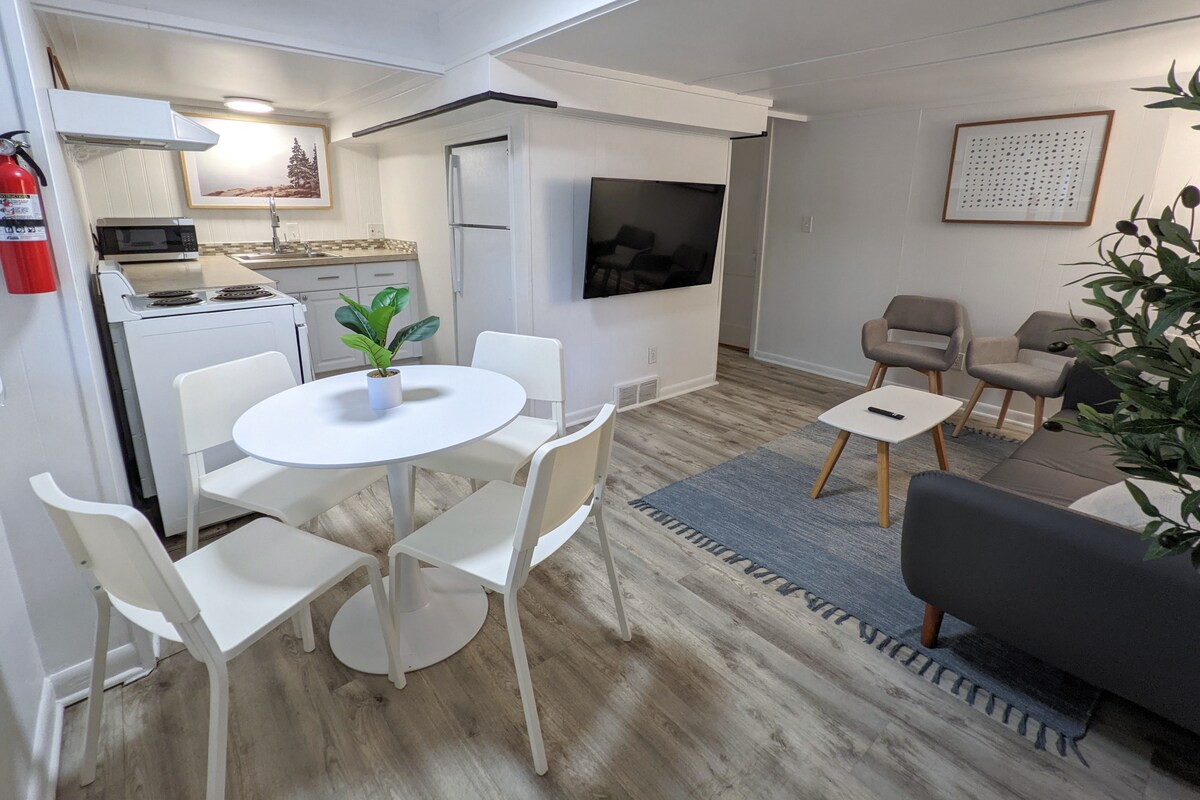 A modern living space features a circular white dining table surrounded by four chairs, with a small green plant at the center. A gray couch and two armchairs create a cozy seating area, while a television is mounted on the wall. Light wood flooring enhances the open layout.