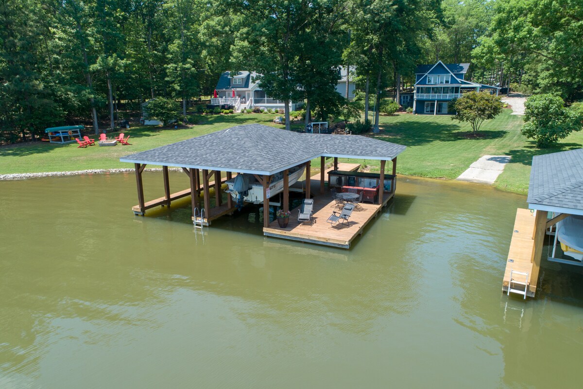 A spacious dock extends over the water, featuring a large covered area with seating and a bar equipped with a mini fridge and sink. Surrounding lush greenery and neighboring cottages create a serene lakeside setting.