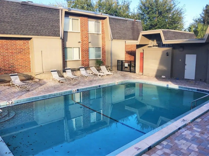 A pool area features a spacious swimming pool surrounded by a paved deck. Several lounge chairs are positioned around the pool, providing seating options. The surrounding building displays a combination of brick and modern siding under a clear sky, contributing to a relaxed atmosphere.