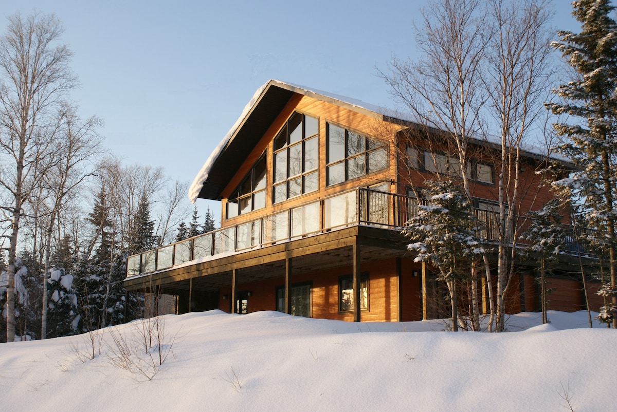 A modern chalet is seen nestled among trees, featuring large glass windows that reflect the snowy landscape. A spacious deck runs along the front, providing outdoor space. The roof is adorned with a layer of fresh snow, adding to the winter ambiance.