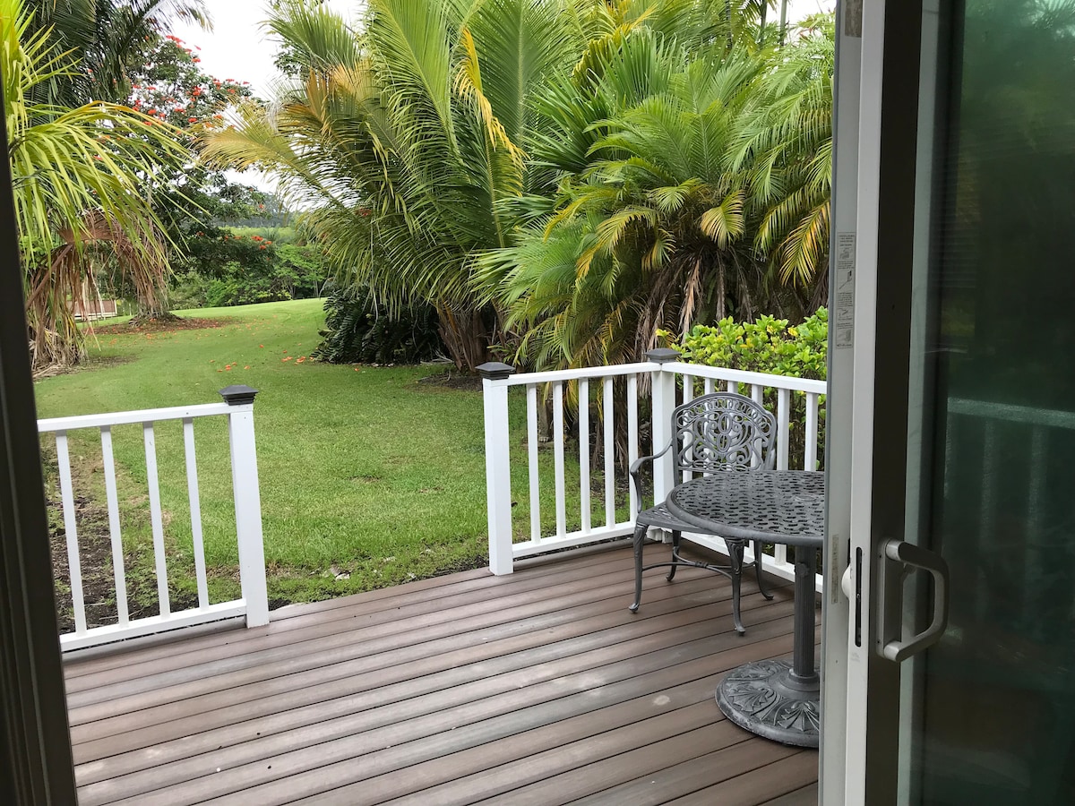 A private patio is visible, featuring a round table and a wrought iron chair. Lush tropical plants surround the area, creating a serene outdoor space that opens onto a grassy lawn. The wooden decking provides a warm and inviting entrance to the patio.