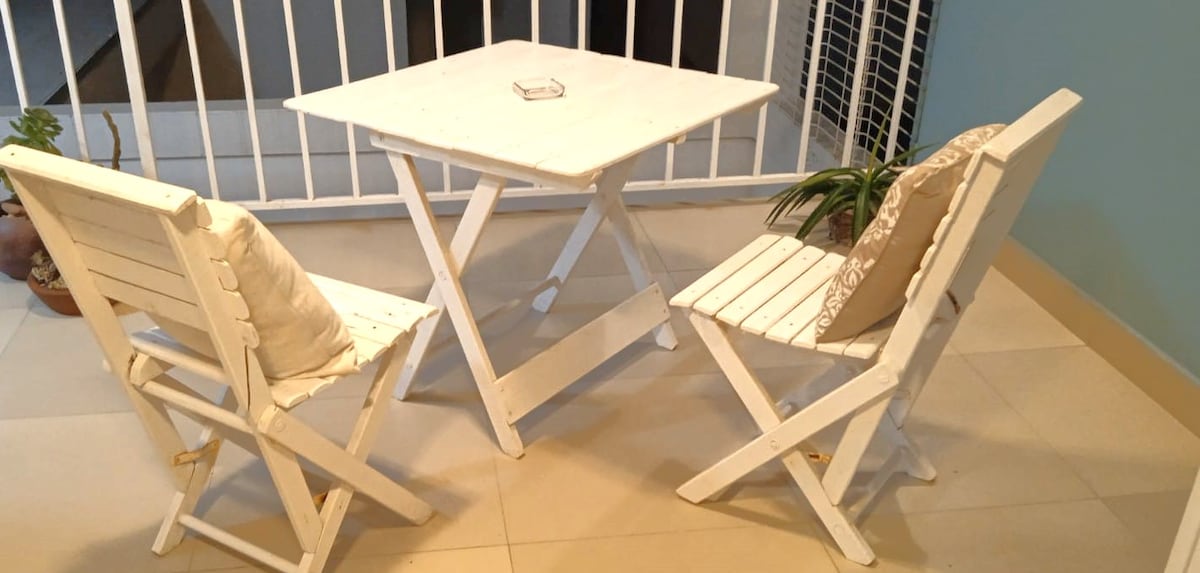 A small outdoor seating area is featured, consisting of a white folding table and two matching chairs. Each chair is adorned with a cushion. A potted plant adds a touch of greenery, while a railing provides a sense of enclosure.
