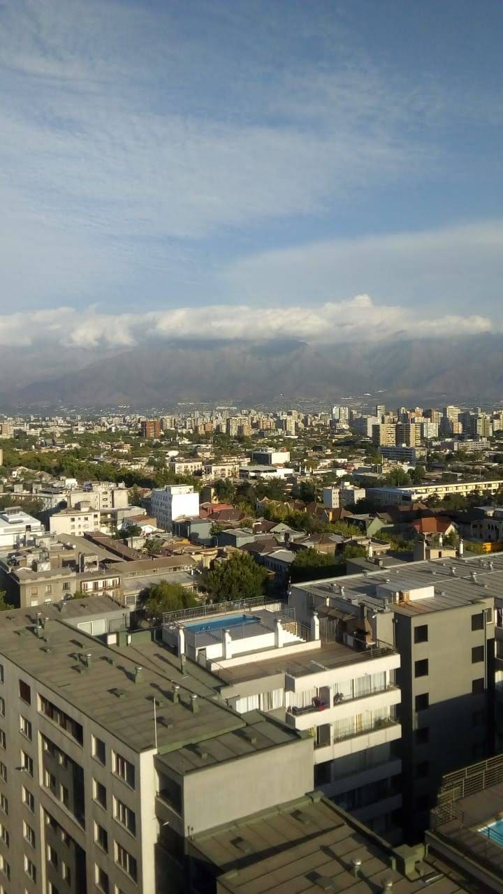 A panoramic view of the city skyline is displayed, featuring a blend of modern buildings and traditional houses. Rolling mountains are seen in the background, under a mostly blue sky with scattered clouds. The diversity of architecture in the urban landscape is noticeable.