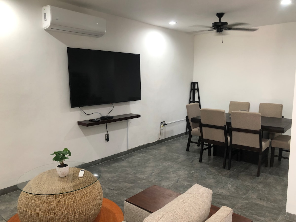 A neutral-colored living area is equipped with a large flat-screen television mounted on the wall. A wooden dining table with six chairs is positioned adjacent to the living space. A small decorative plant rests on a round glass table, complemented by a cozy seating area with light-toned furniture.