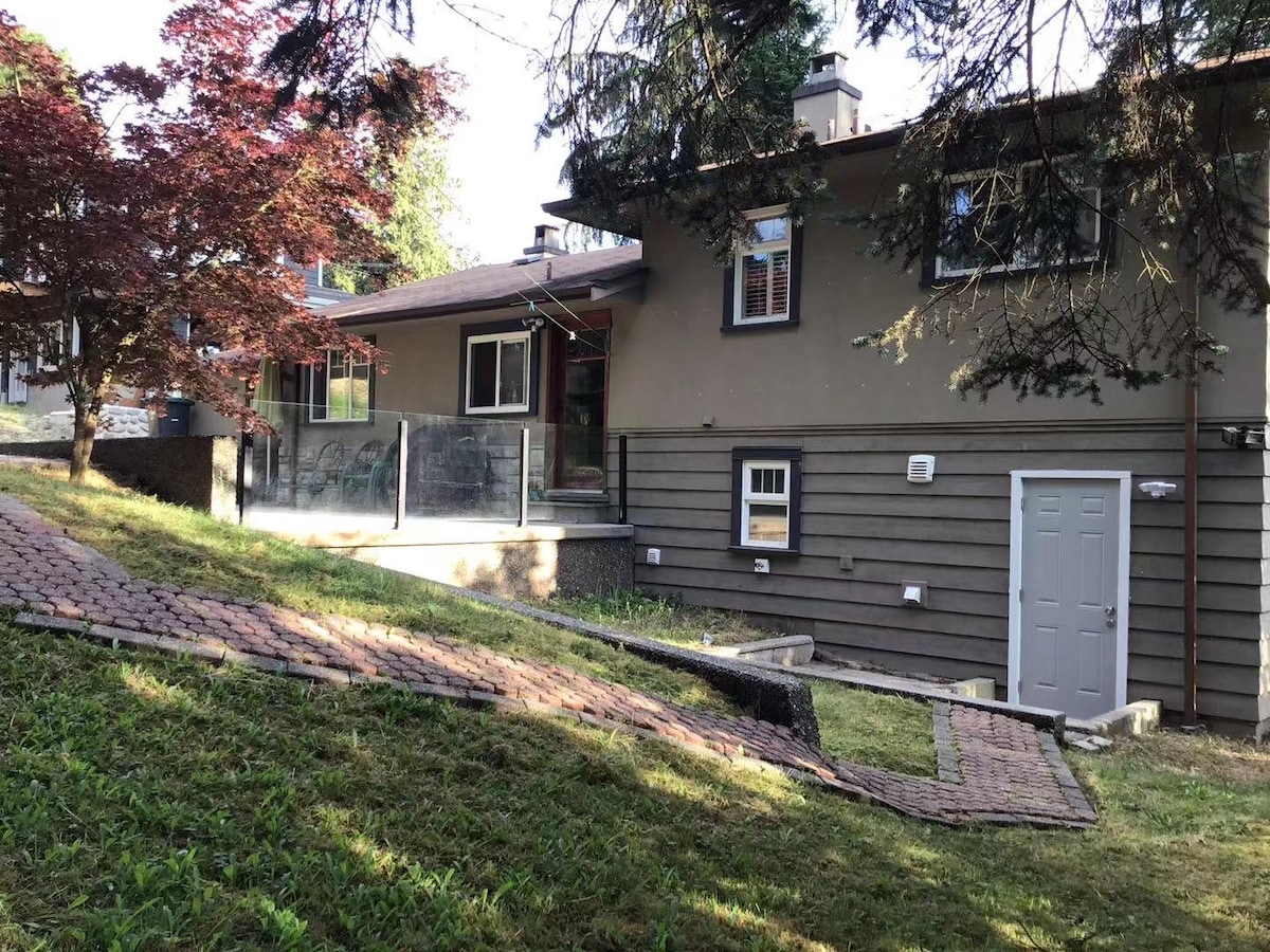 The exterior of a renovated suite is shown, featuring a spacious patio with glass railings. A wooden door is visible along the side of the building, with a landscaped area that includes grass and a path made of stones, highlighted by nearby trees.