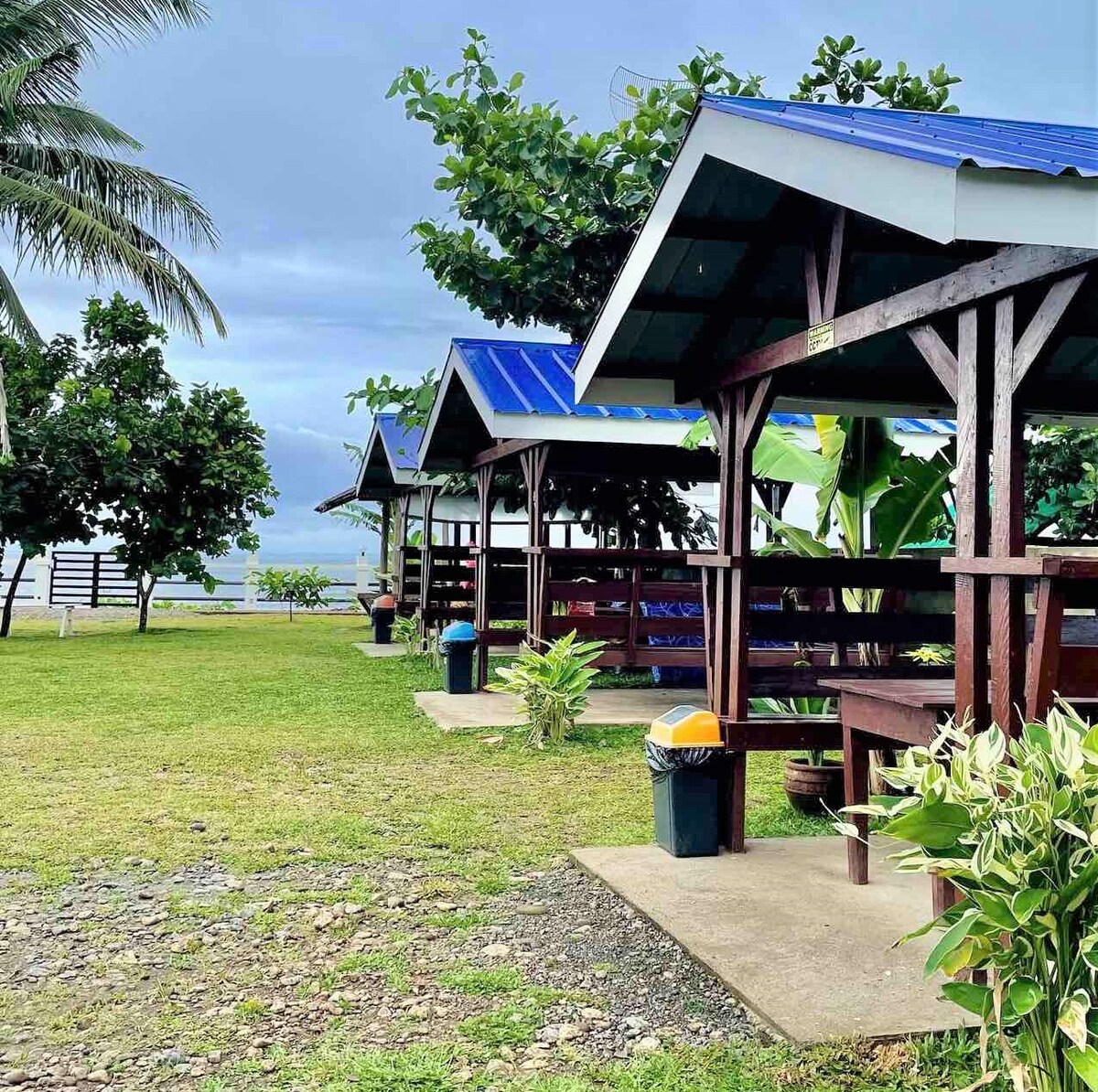 Open huts with blue roofs are positioned on a grassy area, surrounded by lush tropical plants and trees. Trash bins are placed near the huts, and a glimpse of the ocean is visible in the background, enhancing the inviting outdoor setting.