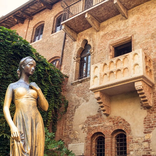 A statue stands in front of a historical building featuring textured brick walls and arched windows. A decorative balcony extends from the upper level, framed by climbing greenery. The setting provides a glimpse into the architectural details and character of the area.
