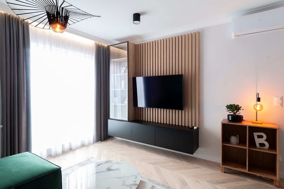A modern living area features a wall-mounted TV above a sleek dark media console. Natural light filters through sheer curtains, illuminating the space. A decorative wooden accent wall adds texture, while a cozy corner lamp and small indoor plant enhance the ambiance.
