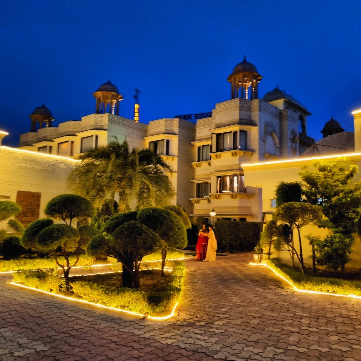 The exterior of the resort is lit by warm lights, surrounded by well-manicured shrubs and palm trees. Elegant architecture features traditional dome shapes. Two guests are seen walking along a paved path, creating a serene atmosphere at twilight.